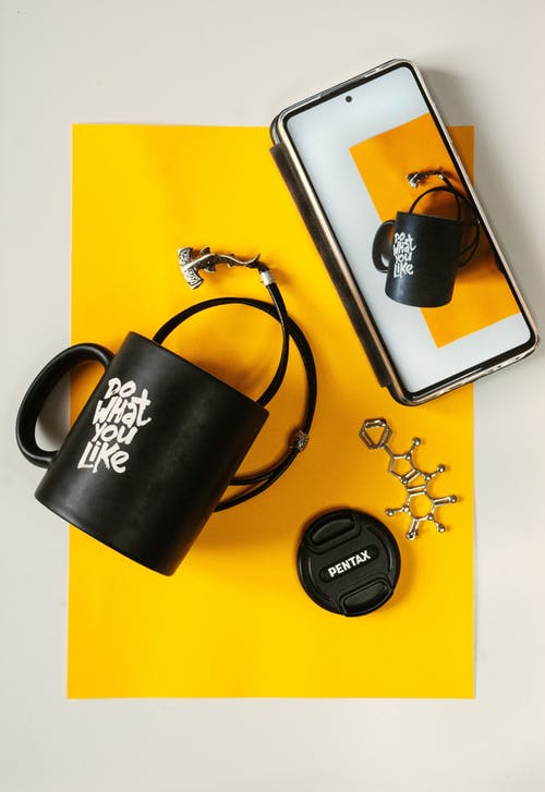 Free From above of creative composition of mug and smartphone with similar photo on screen placed on yellow paper with accessories on white desk Stock Photo